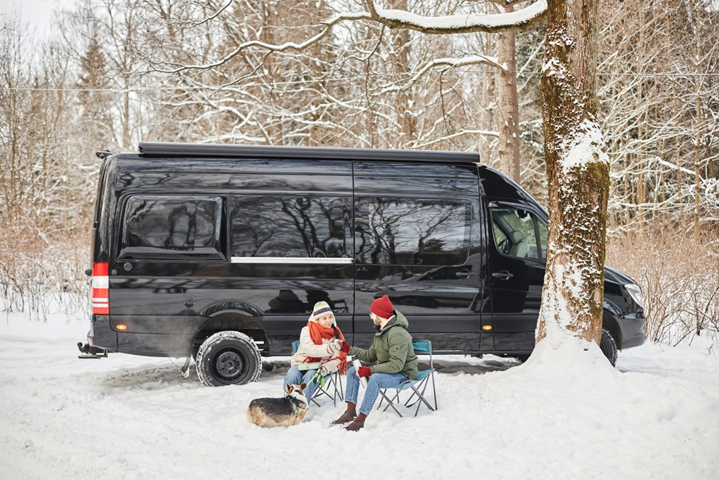A young couple sits outside of their camper van with their dog on a snowy winter day.