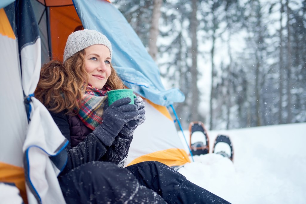 A woman looking from her tent and enjoying a warm drink during a winter camping trip.