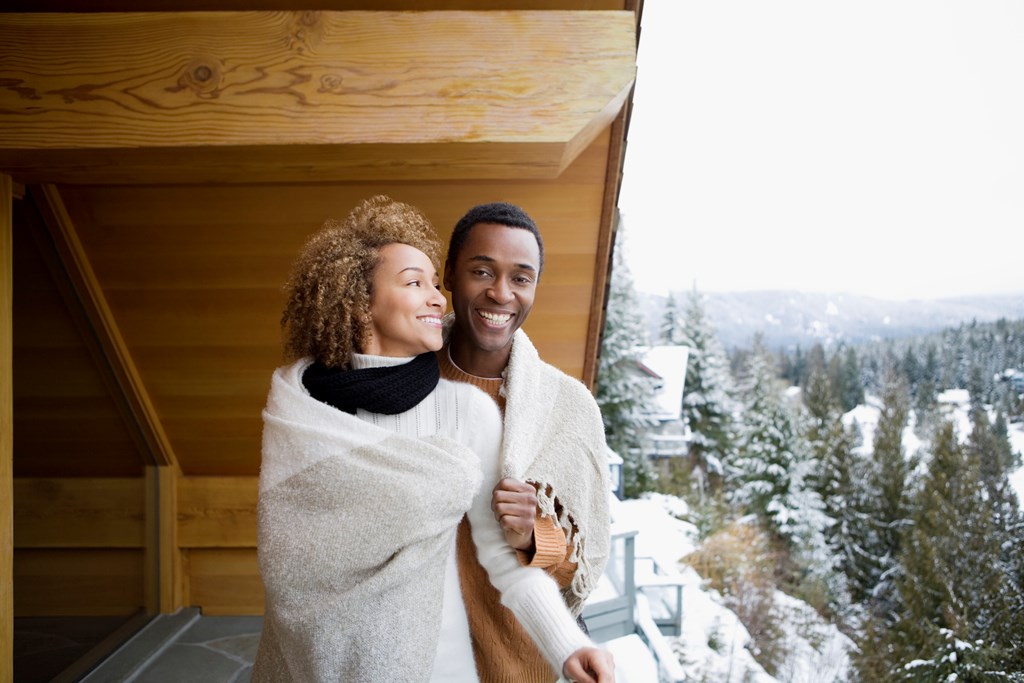 A happy couple wrapped in a blanket looks out at a snowy scene from the deck of a cabin in winter.