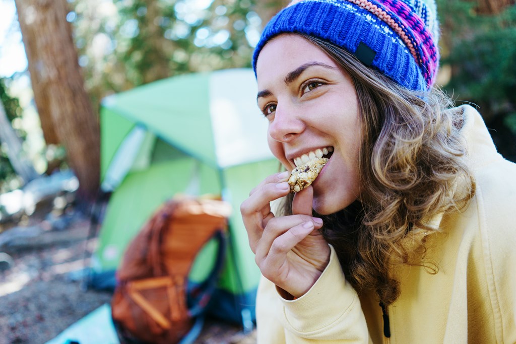 A young woman in a winter hat eats a granola bar in front of her green tent.
