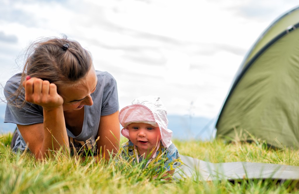 A mother and her infant son sit in the grass in front of their camping tent.