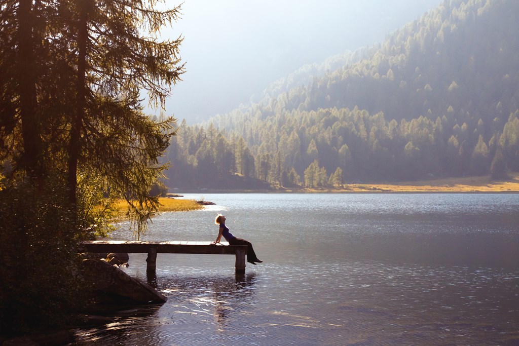 A young woman sits at the end of dock on a mountain lake.