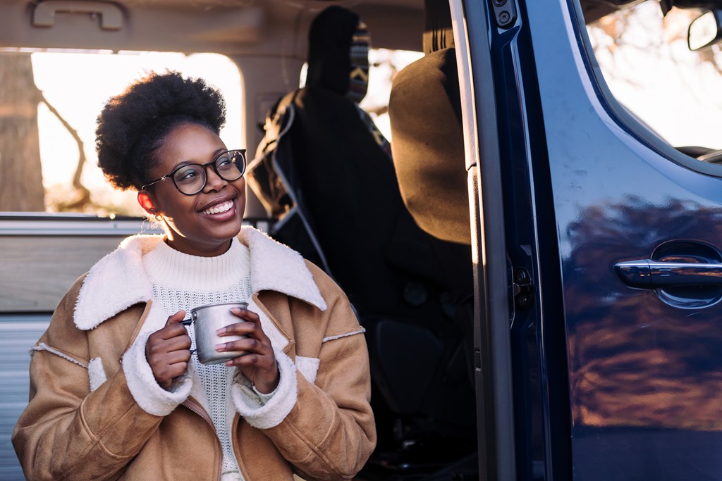 A young Black woman drinks coffee during a chilly morning in front of her camper van.