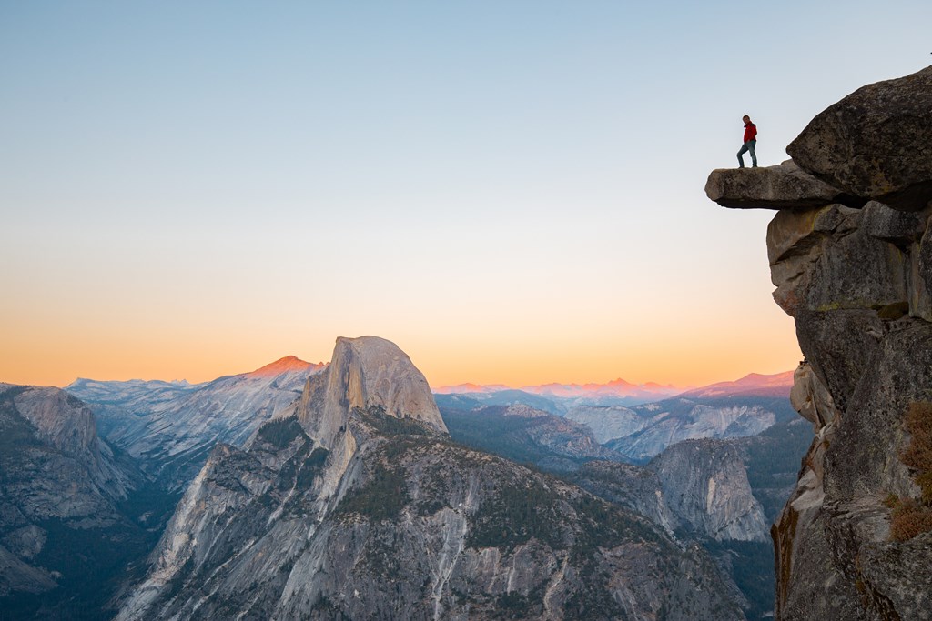 A fearless hiker is standing on an overhanging rock enjoying the view towards famous Half Dome at Glacier Point overlook in beautiful evening twilight, Yosemite National Park, California, USA