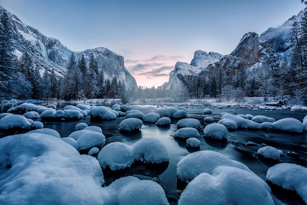 Snow covers rocks and trees in the Yosemite Valley just before sunrise.