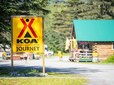 A large yellow and brown Valdez KOA Journey campground sign, featuring the KOA logo, standing near lush green trees and path under a bright sky.
